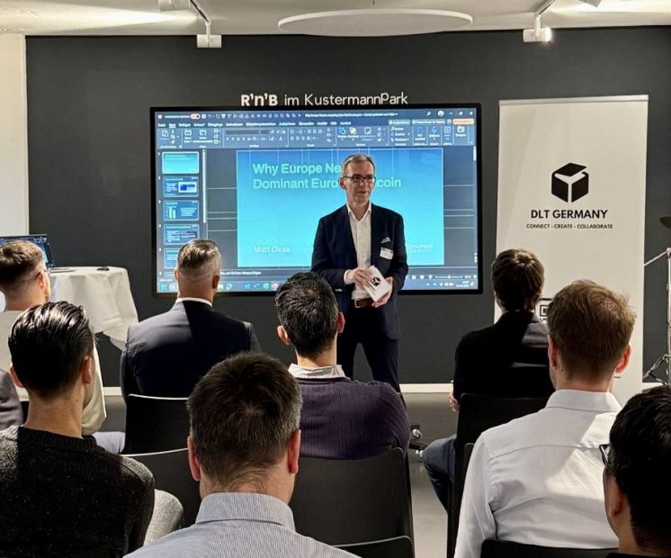Ein Mann in Geschäftskleidung hält vor einem Publikum einen Vortrag mit dem Titel "Warum Europa eine dominante Euro-Münze braucht", der sich auf Innovationen konzentriert. Im Hintergrund sind ein großer Bildschirm und ein DLT Germany-Banner zu sehen.