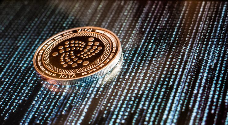 Eine physische IOTA-Münze ruht auf einer Oberfläche, die einen digitalen Binärcode und alphanumerische Zeichen anzeigt.