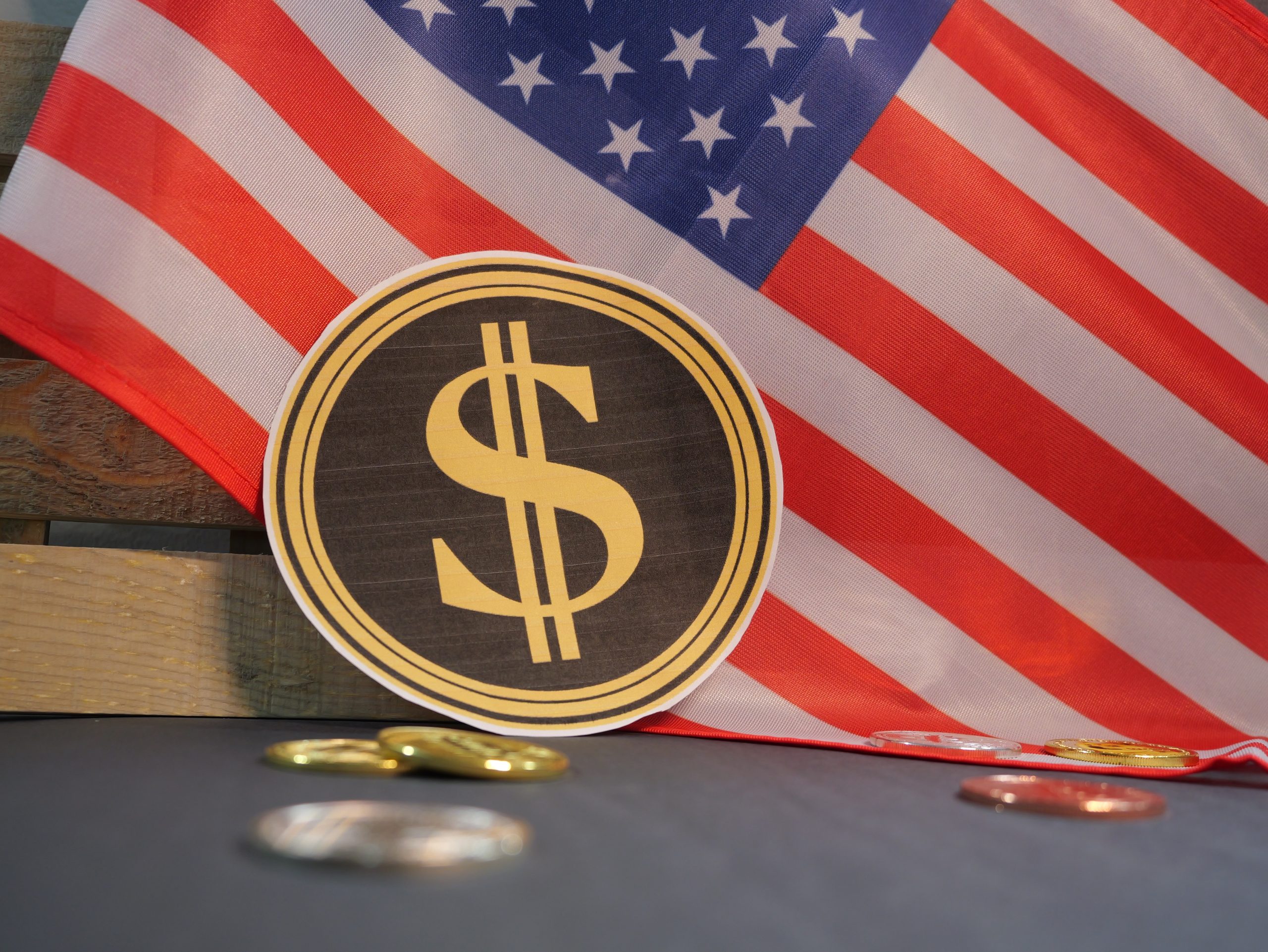 Ein goldenes Dollar-Symbol ist vor einer Flagge der Vereinigten Staaten platziert, mit verstreuten Münzen auf einer dunklen Oberfläche, die an das Konzept von Stablecoin und digitaler Währung erinnert.