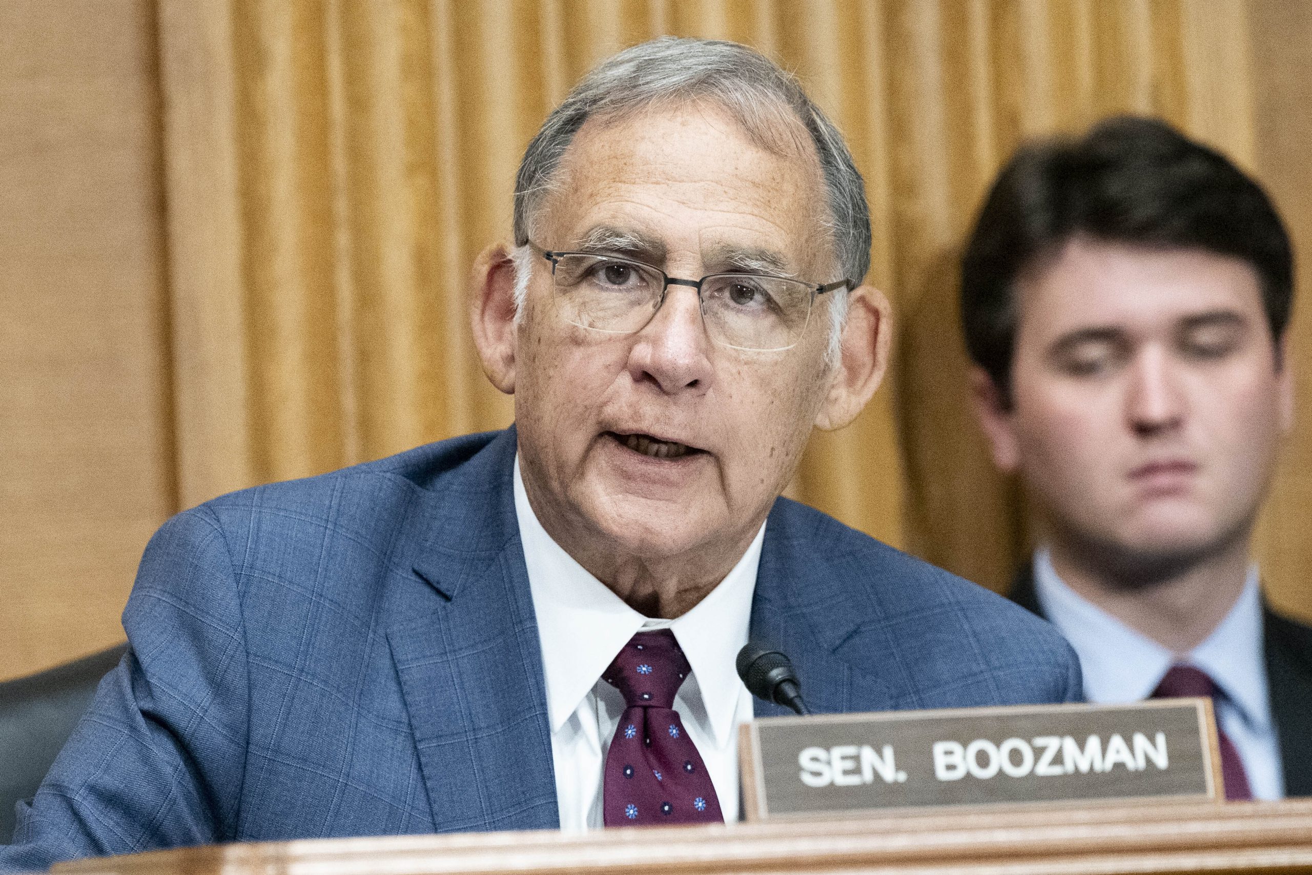 Ein älterer Mann in Anzug und Krawatte spricht während einer formellen Sitzung zu einem Mikrofon, vor ihm ein Namensschild mit der Aufschrift Sen. Boozman. Eine weitere Person sitzt im Hintergrund.