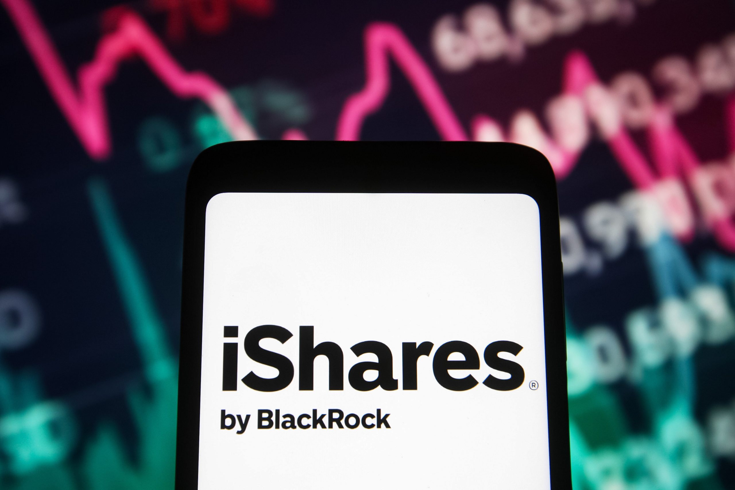 Auf einem Smartphone ist das iShares by BlackRock-Logo zu sehen, mit einem unscharfen Hintergrund, der eine Grafik mit Börsendaten und Bitcoin-Trends zeigt, die durch rosa und grüne Linien hervorgehoben werden.