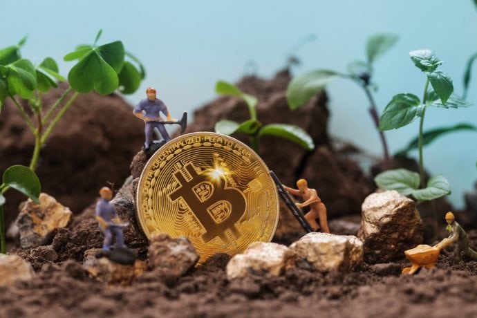 Miniaturfiguren, die Bitcoin-Mining betreiben, umgeben eine große Bitcoin-Münze und graben sich durch Erde und Goldnuggets inmitten üppiger grüner Pflanzen.