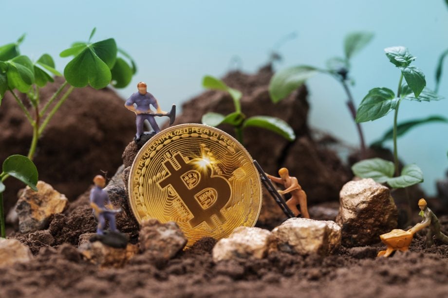 Miniaturfiguren, die Bitcoin-Mining betreiben, umgeben eine große Bitcoin-Münze und graben sich durch Erde und Goldnuggets inmitten üppiger grüner Pflanzen.