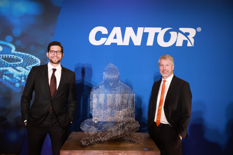 Zwei Männer in Anzügen, darunter Michael Saylor, stehen zu beiden Seiten einer modernen abstrakten Skulptur vor einer blauen Wand, auf der das Wort CANTOR prangt.