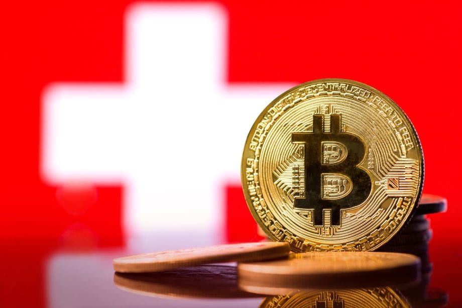 Eine goldene Bitcoin-Münze steht aufrecht vor einer Schweizer Flagge.