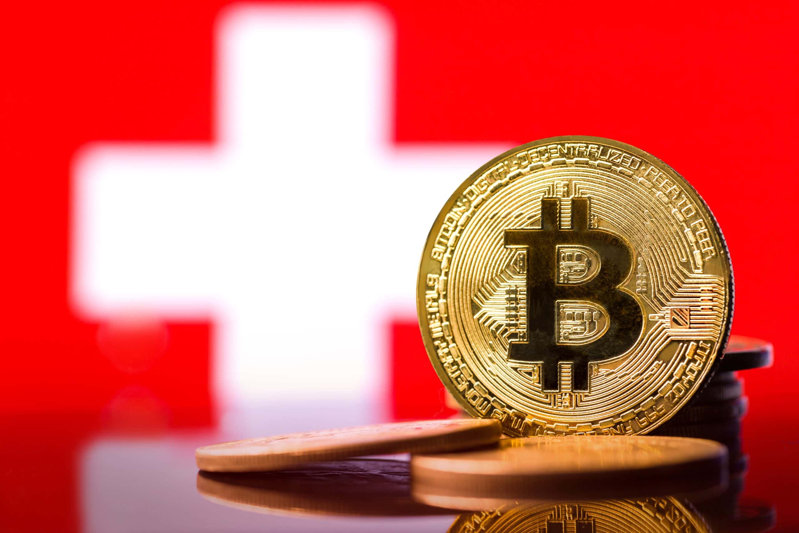Eine goldene Bitcoin-Münze steht aufrecht vor einer Schweizer Flagge.