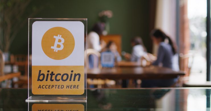 Auf einem Schild steht "bitcoin accepted here", dahinter sind Menschen zu sehen