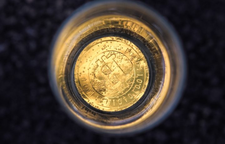 Eine goldene Bitcoin-Münze, von oben gesehen, eingetaucht in ein klares Glas, das mit einer hellen Flüssigkeit und kleinen Blasen gefüllt ist, unterstreicht die Faszination von Bitcoin vor einem dunklen, unscharfen Hintergrund.