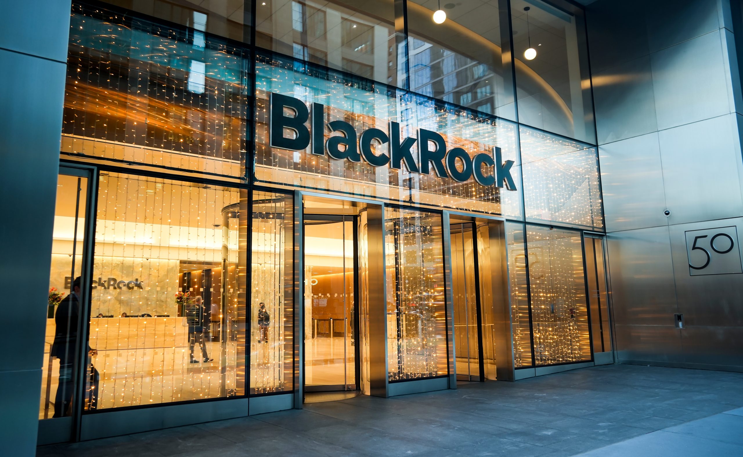 Eingang zu einem modernen Bürogebäude mit großen Glastüren und einem Schild mit der Aufschrift BlackRock über dem Eingang, beleuchtet von Innenleuchten - ein Zentrum für Gespräche über Investitionen und Kryptowährungen.