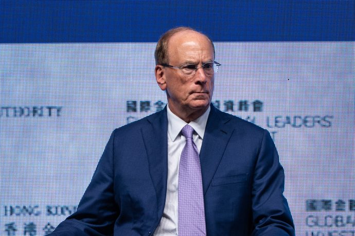 Ein Mann im blauen Anzug und mit hellvioletter Krawatte sitzt vor einem Bildschirm, auf dem ein Text über Bitcoin angezeigt wird, was wie eine formelle Veranstaltung oder Konferenz aussieht.