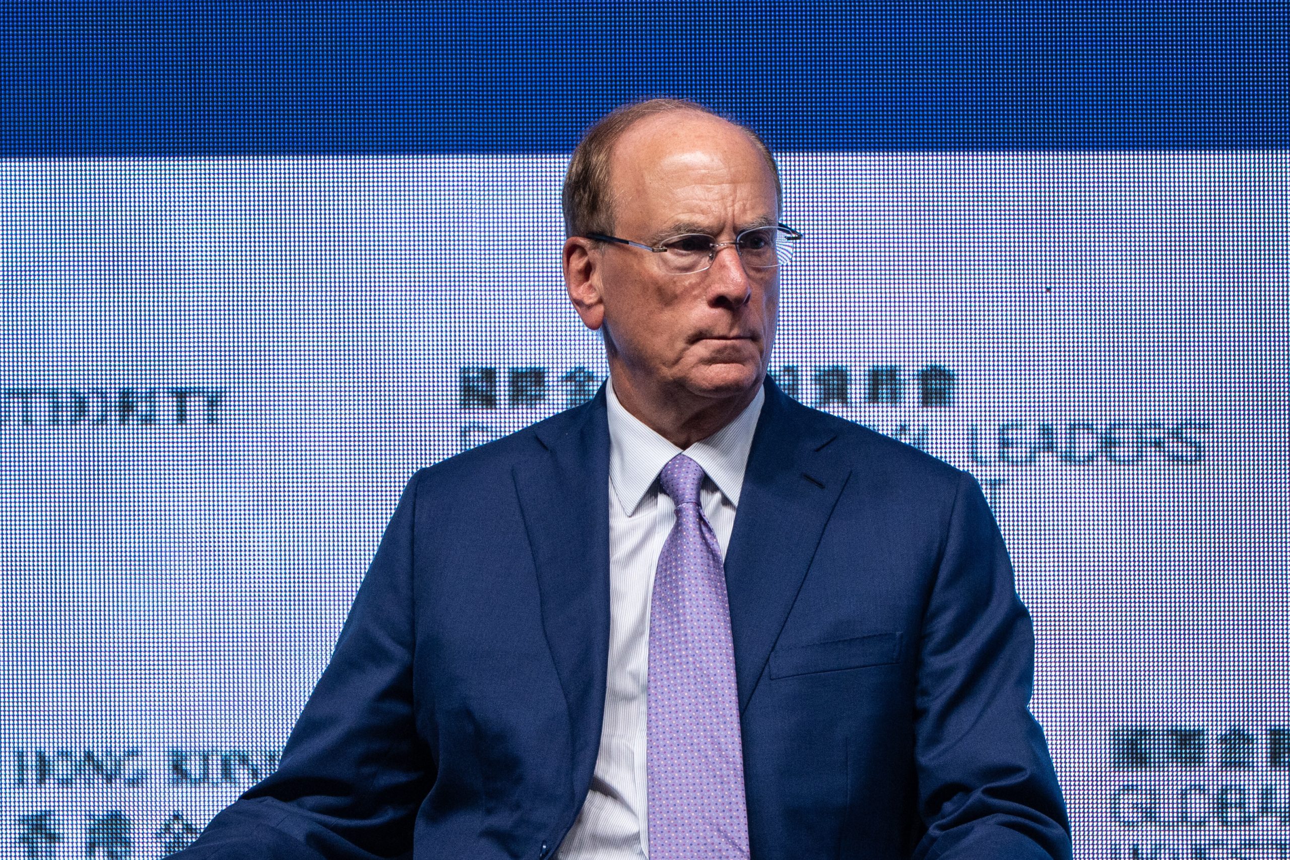 Ein Mann im blauen Anzug und mit hellvioletter Krawatte sitzt vor einem Bildschirm, auf dem ein Text über Bitcoin angezeigt wird, was wie eine formelle Veranstaltung oder Konferenz aussieht.