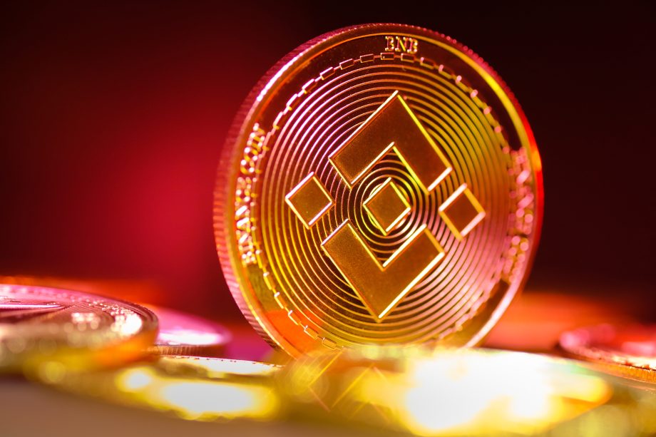 Eine Nahaufnahme einer aufrecht stehenden Binance-Goldmünze, auf der das Binance-Logo vor einem verschwommenen roten und gelben Hintergrund zu sehen ist.