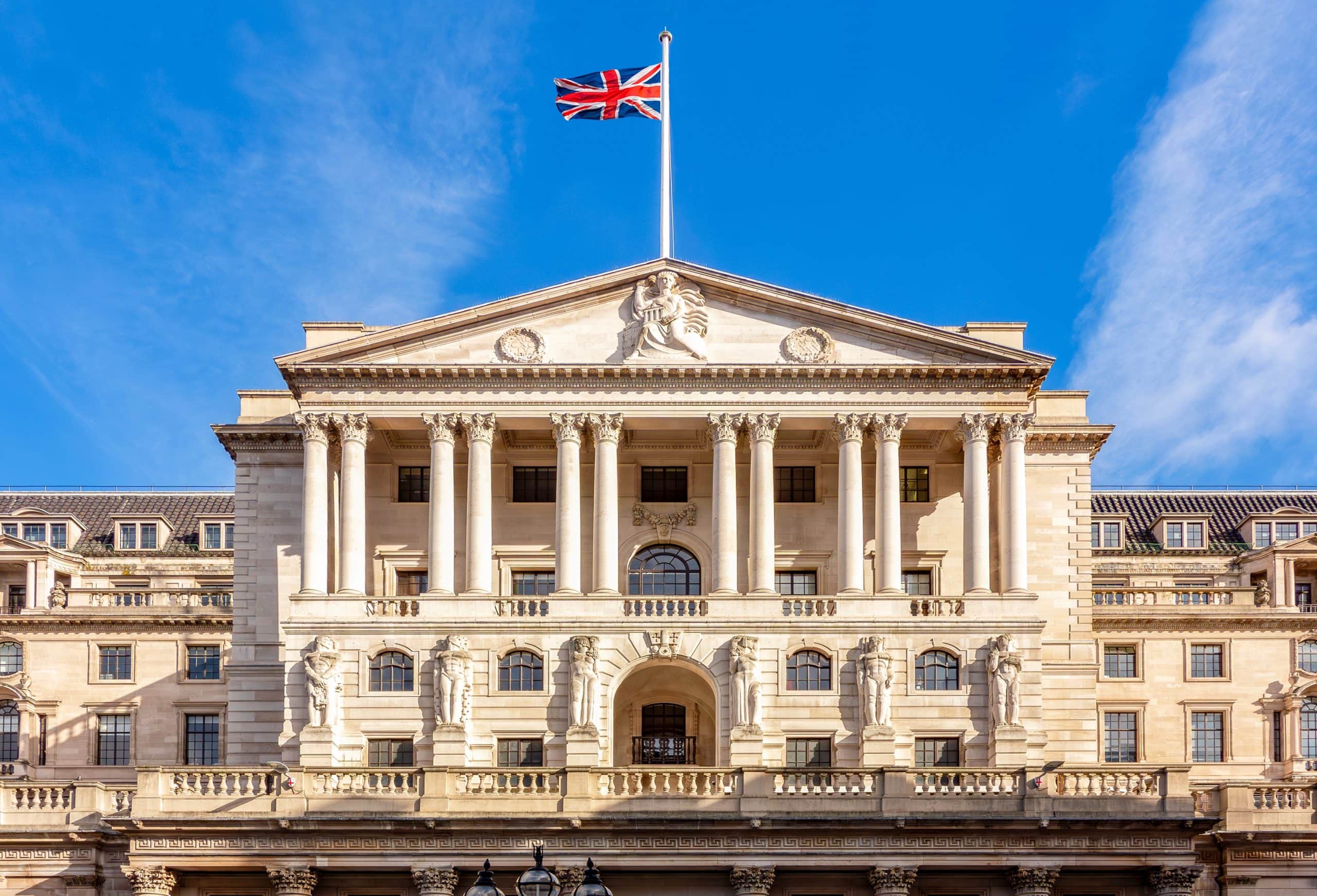 Die Fassade des Gebäudes der Bank of England mit Säulen, Steinskulpturen und einer britischen Flagge an der Spitze unter einem strahlend blauen Himmel - ein Symbol für Tradition und Innovation im Finanzwesen, von Banknoten bis hin zu Diskussionen über stabile Münzen.