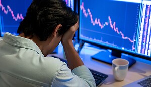 Eine Person sitzt an einem Schreibtisch mit dem Kopf in den Händen und blickt auf Computerbildschirme, die abnehmende Aktiencharts anzeigen, darunter auch die Performance von Cardano.