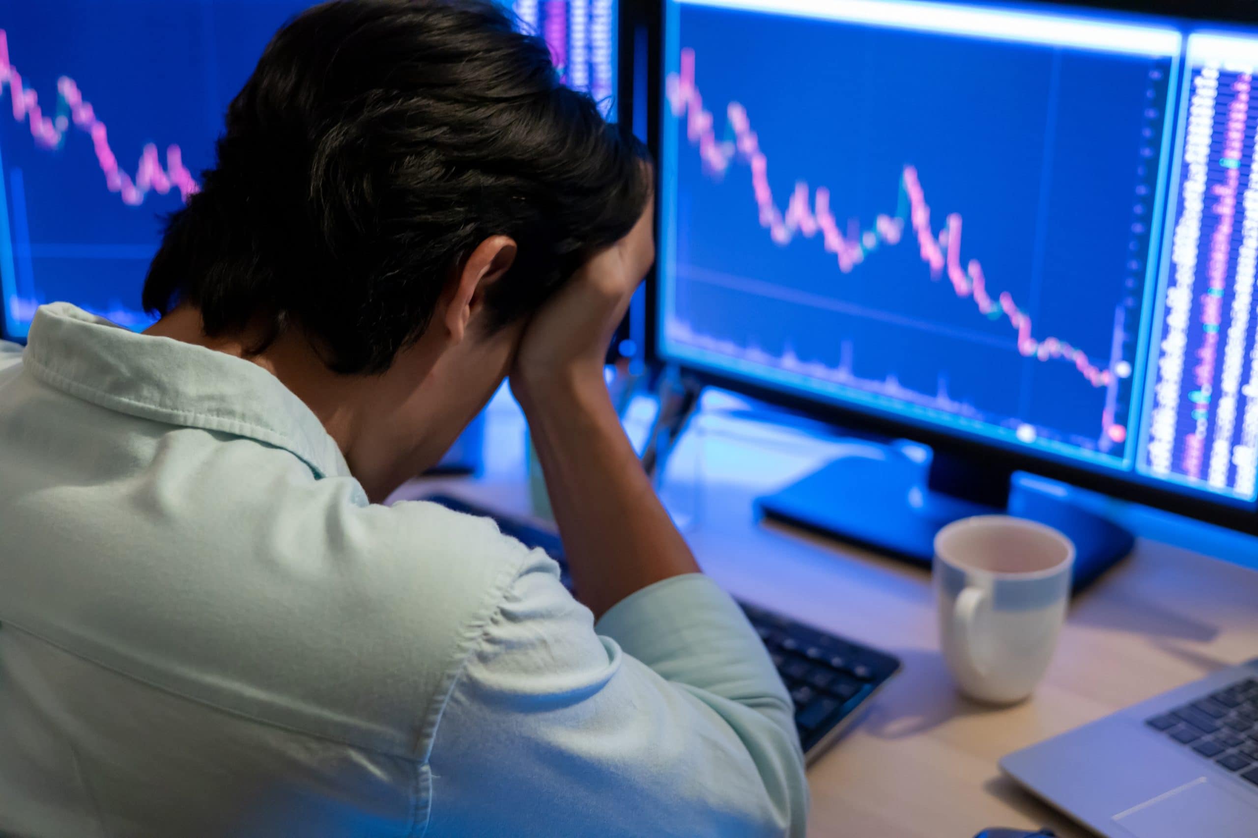 Eine Person sitzt an einem Schreibtisch mit dem Kopf in den Händen und blickt auf Computerbildschirme, die abnehmende Aktiencharts anzeigen, darunter auch die Performance von Cardano.