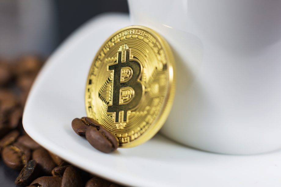 Eine goldene Bitcoin-Münze lehnt an einer weißen Tasse mit Untertasse, in deren Nähe Kaffeebohnen verstreut sind - eine stilvolle Anspielung auf die Welt von Krypto und Kaffeekultur.