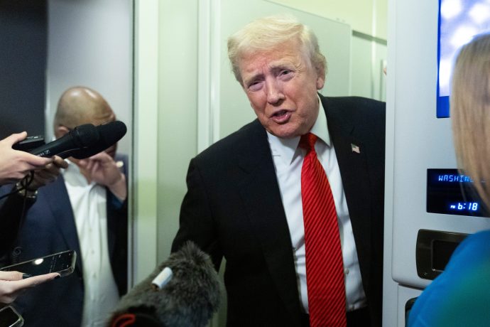 Ein Mann im Anzug und mit roter Krawatte (Donald Trump) spricht mit Reportern, die Mikrofone vor sich haben, in etwas, das wie ein Flugzeug oder ein enger Gang aussieht.