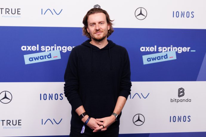 Ein Mann mit schwarzem Kapuzenpulli (Eric Demuth) steht vor einem Step-and-Repeat-Banner mit den Logos von Axel Springer Award, Mercedes-Benz, Bitpanda, IAV, IONOS und anderen Sponsoren.