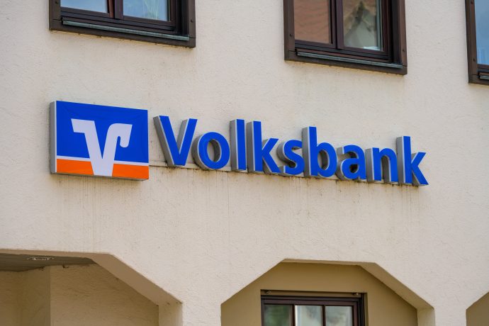 Die Außenseite eines Gebäudes mit einem blau-orangefarbenen Volksbank-Schild, das die Einhaltung der MiCA-Richtlinien widerspiegelt, ist an der Wand unter zwei Fenstern angebracht.