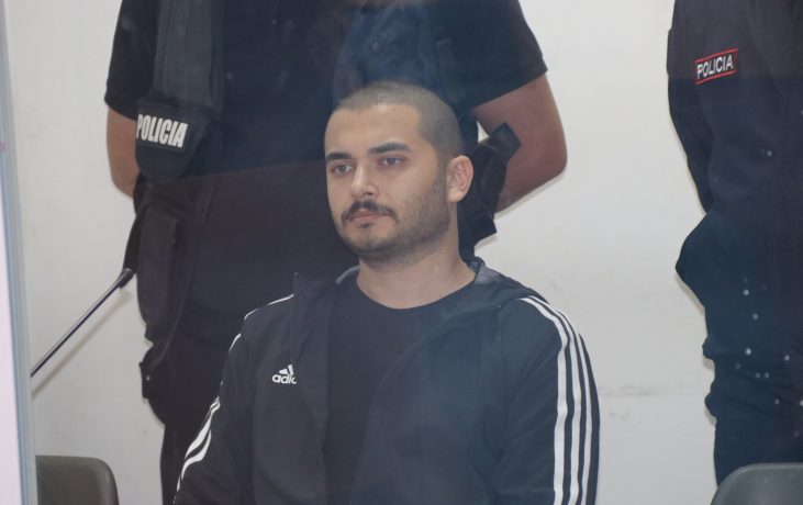 Ein Mann in einem schwarzen Trainingsanzug mit weißen Streifen, der mit Thodex in Verbindung stehen soll, sitzt vor zwei uniformierten Polizeibeamten vor einem weißen Hintergrund.