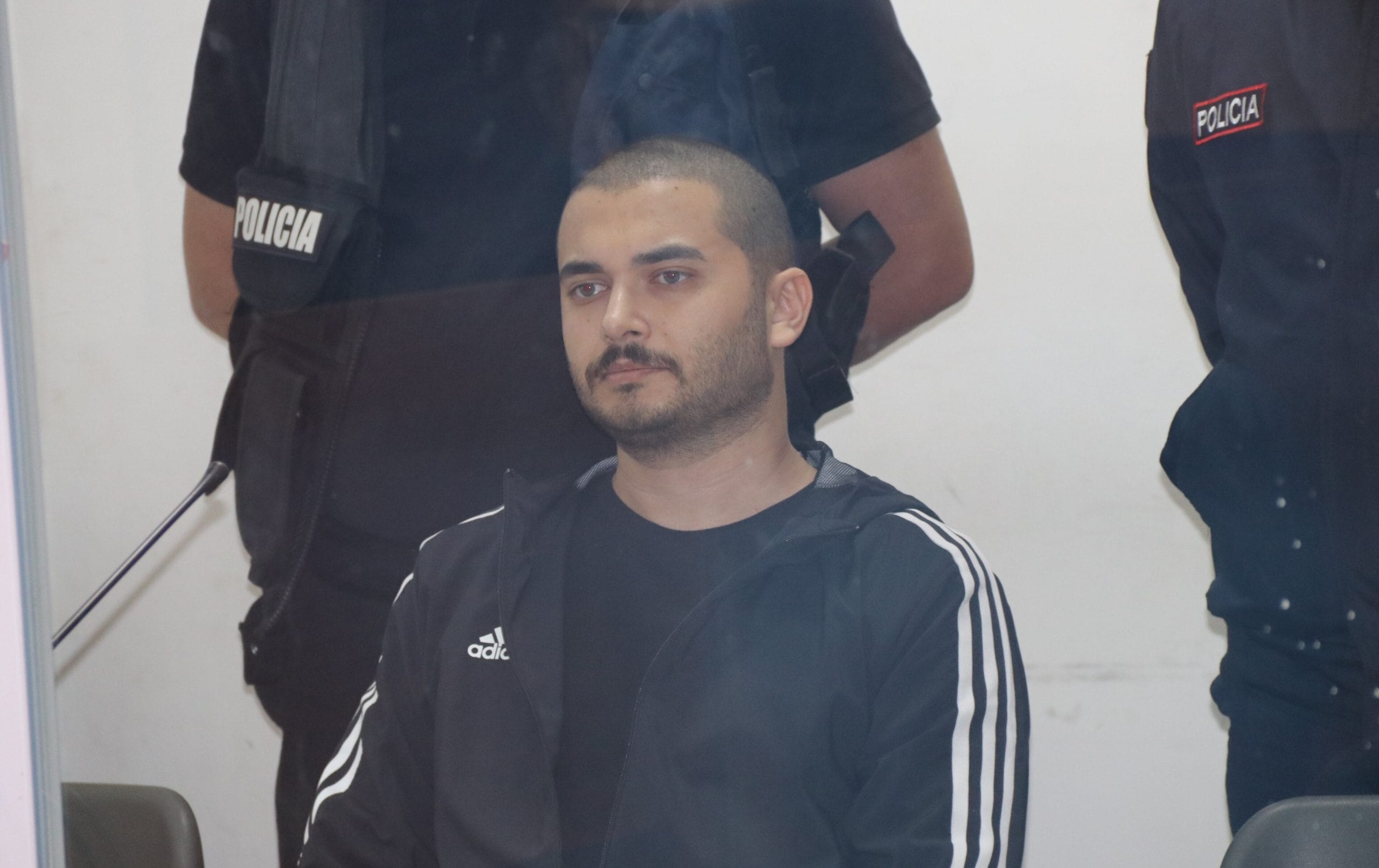 Ein Mann in einem schwarzen Trainingsanzug mit weißen Streifen, der mit Thodex in Verbindung stehen soll, sitzt vor zwei uniformierten Polizeibeamten vor einem weißen Hintergrund.