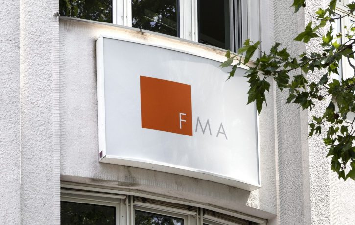 Eine weiße Gebäudefassade zeigt ein rechteckiges Schild mit einem orangefarbenen Quadrat und den Buchstaben FMA daneben, die auf eine moderne Krypto-Präsenz hindeuten. Im Vordergrund sind Baumzweige zu sehen.