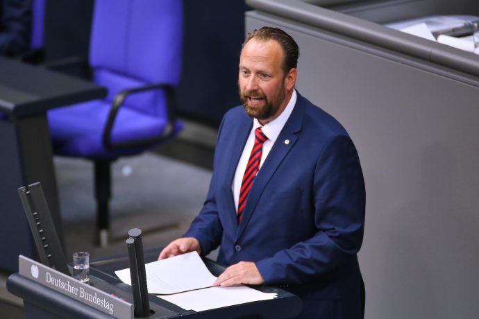 Ein Mann im blauen Anzug und mit rot gestreifter Krawatte spricht an einem Podium mit der Aufschrift Deutscher Bundestag in einer formellen Umgebung, hält Papiere in der Hand und spricht möglicherweise über Themen wie die Bitcoin-Regulierung.