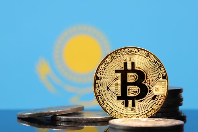 Eine goldene Bitcoin-Münze steht aufrecht zwischen gestapelten Münzen, im Hintergrund ist die Flagge von Kasachstan zu sehen.