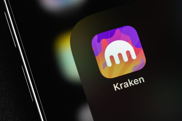 Nahaufnahme eines Smartphone-Bildschirms, auf dem das Kraken-App-Symbol mit dem charakteristischen weißen Kraken-Logo auf einem leuchtenden lila, orangefarbenen und gelben Hintergrund angezeigt wird.