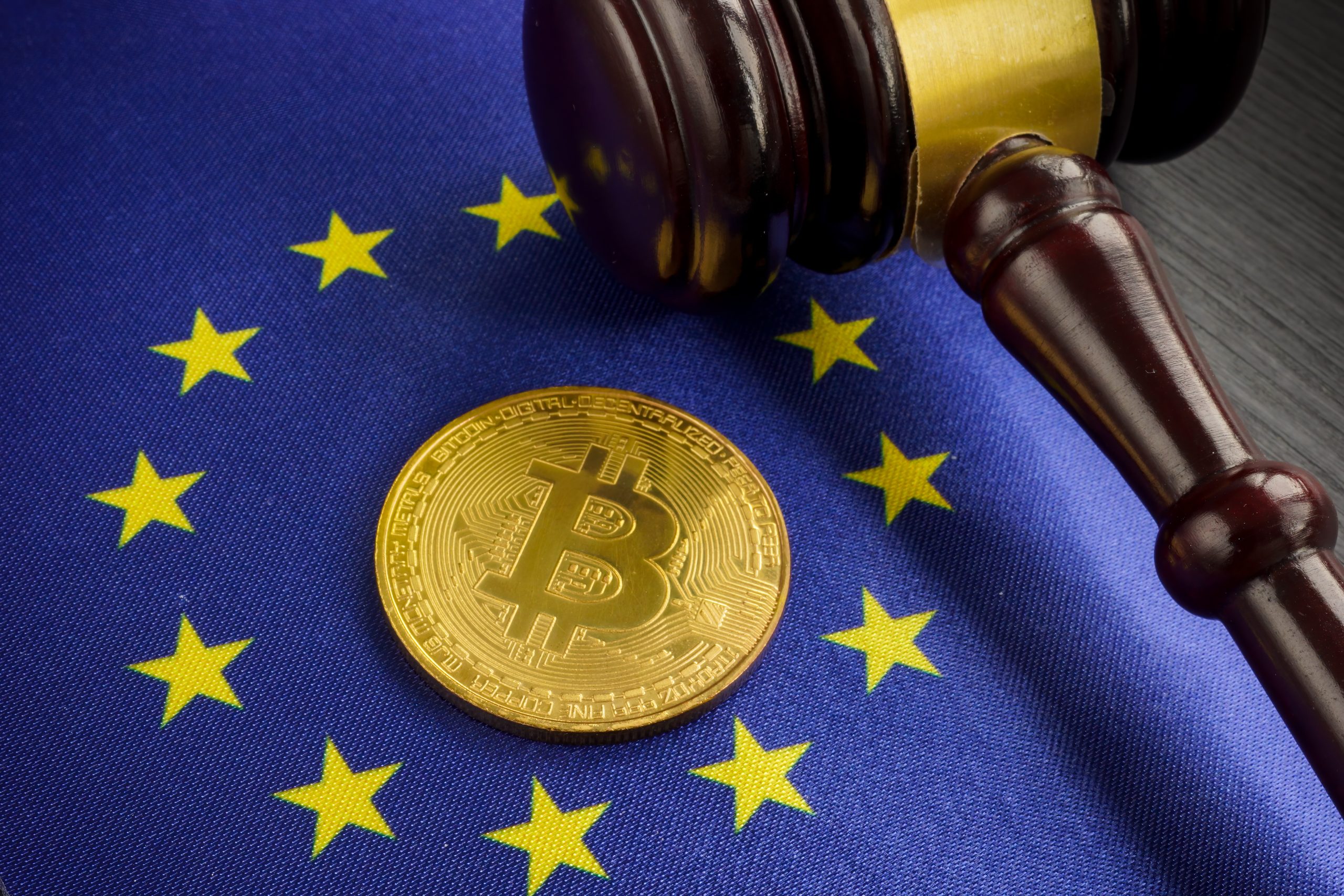 Ein Hammer und eine Bitcoin-Münze liegen auf der Flagge der Europäischen Union und symbolisieren die Krypto-Regulierung in Europa.
