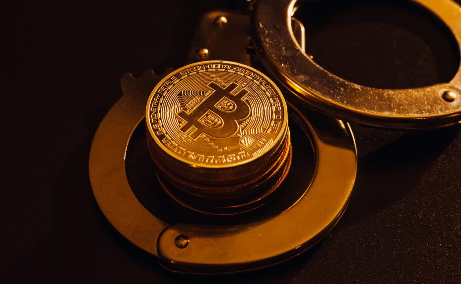 Ein Stapel Bitcoin-Münzen sitzt in einem offenen Paar Metallhandschellen auf einer dunklen Oberfläche und symbolisiert die Spannung zwischen Krypto-Freiheit und Regulierung.