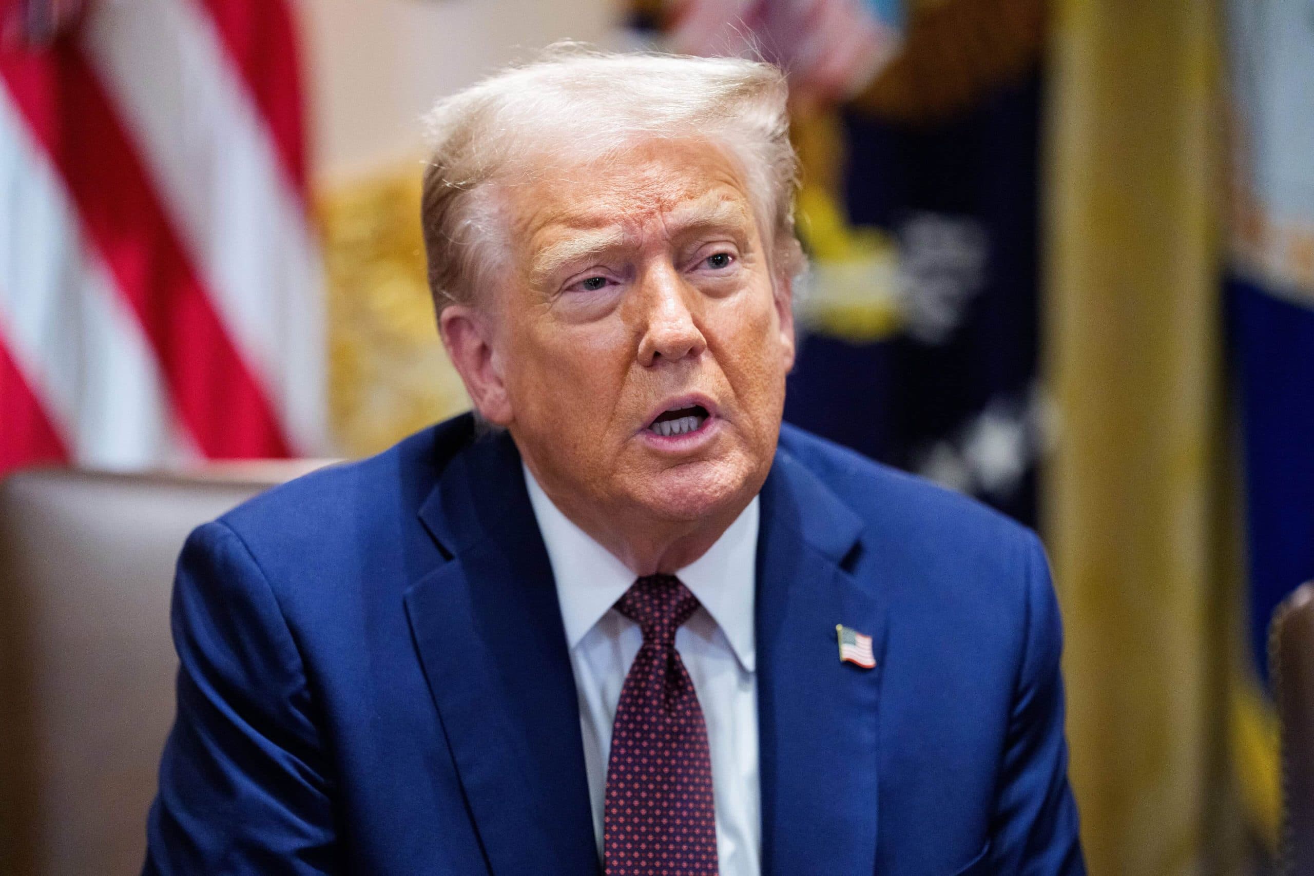 Ein Mann mit heller Haut und blondem Haar, der einen marineblauen Anzug, ein weißes Hemd und eine rote Krawatte mit dem Krypto-Logo trägt, sitzt in einem Haus vor amerikanischen Flaggen.