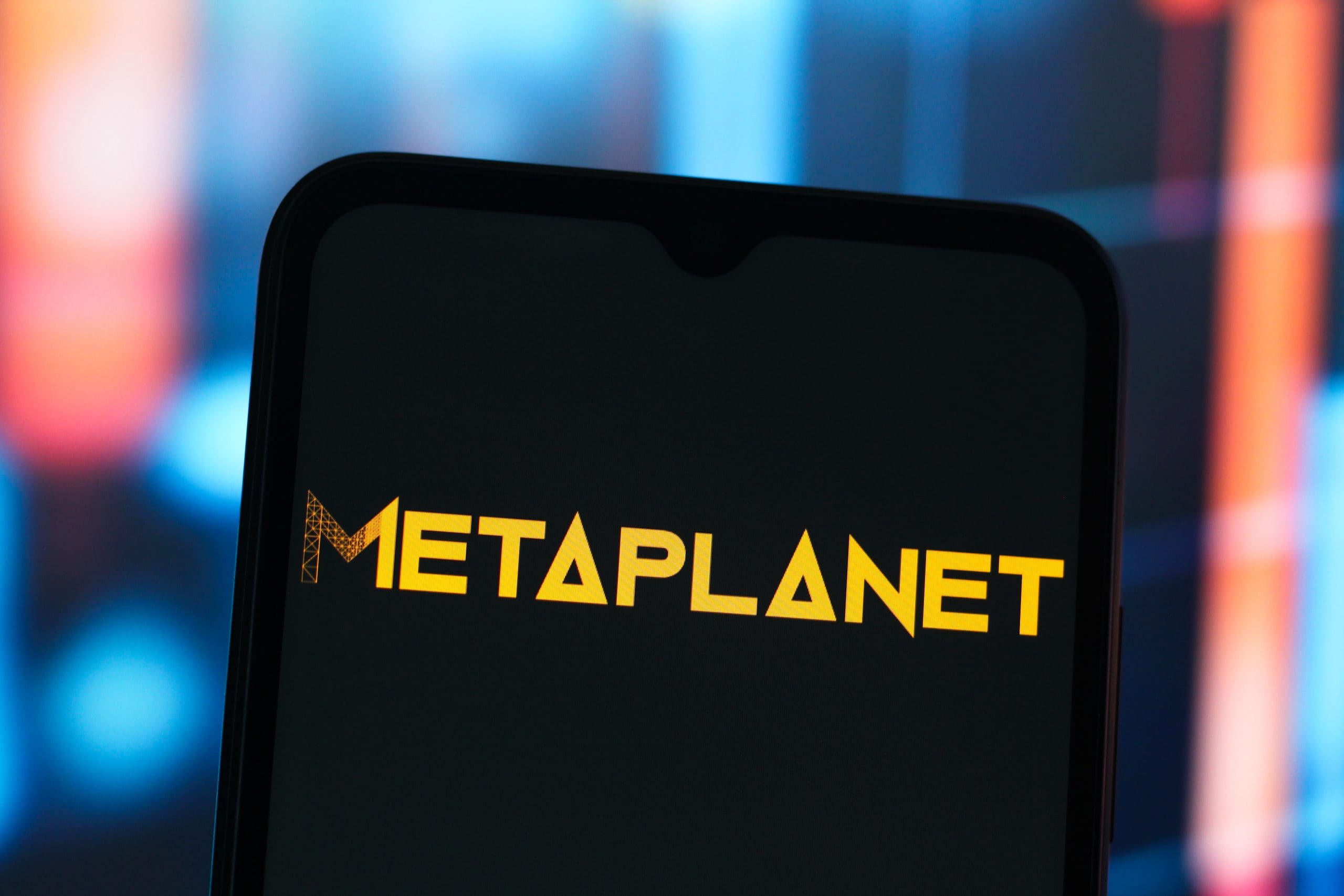 Ein Smartphone-Bildschirm zeigt das Wort METAPLANET in fetten gelben Buchstaben vor einem dunklen Hintergrund mit verschwommenen farbigen Lichtern.