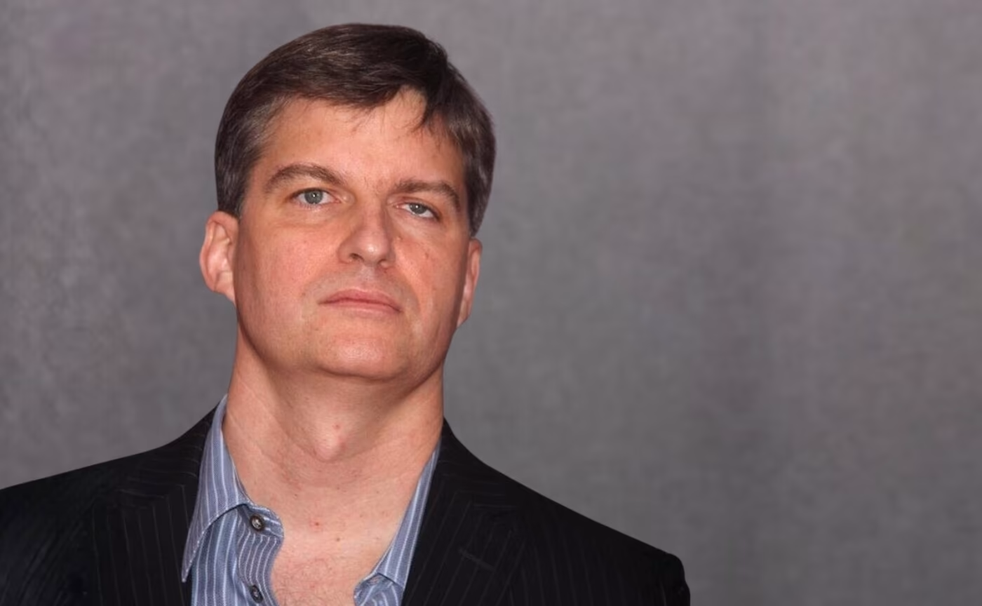 Ein Mann mit kurzen braunen Haaren, der an Michael Burry erinnert, steht in einem dunklen Nadelstreifenanzug vor einem schlichten grauen Hintergrund und schaut direkt in die Kamera - die Intensität eines Mannes, der bereit ist für einen plötzlichen Abgang am Markt.