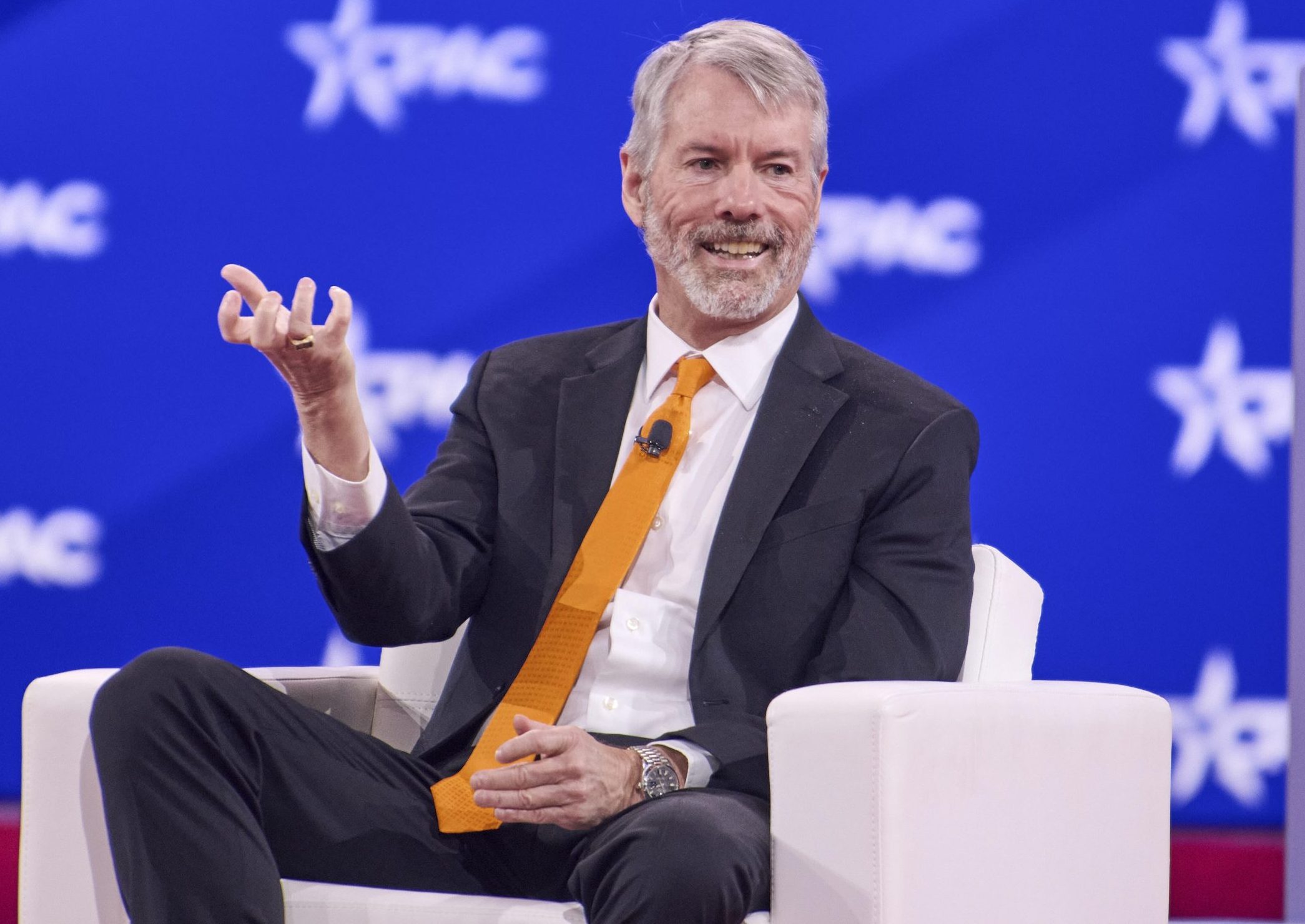 Ein Mann im Anzug und mit orangefarbener Krawatte (Michael Saylor) sitzt auf einem weißen Stuhl und gestikuliert mit seiner Hand während einer Podiumsdiskussion über Bitcoin vor einem blauen Hintergrund mit weißen Sternlogos.