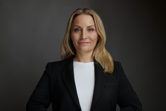 Eine Frau mit langen blonden Haaren, die einen schwarzen Blazer und ein weißes Oberteil trägt, steht vor einem schlichten dunklen Hintergrund und blickt mit neutraler Miene in die Kamera, so ruhig wie das Meer, bevor sich der Krake regt.