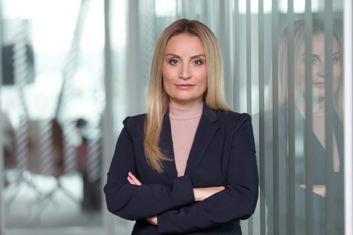 Eine Frau mit langen blonden Haaren in einem dunklen Anzug steht mit verschränkten Armen in einem modernen Büro, ihr Spiegelbild ist auf einer Glaswand neben ihr zu sehen, und sie strahlt die Zuversicht und Präsenz eines Kraken aus, der durch unbekannte Gewässer navigiert.