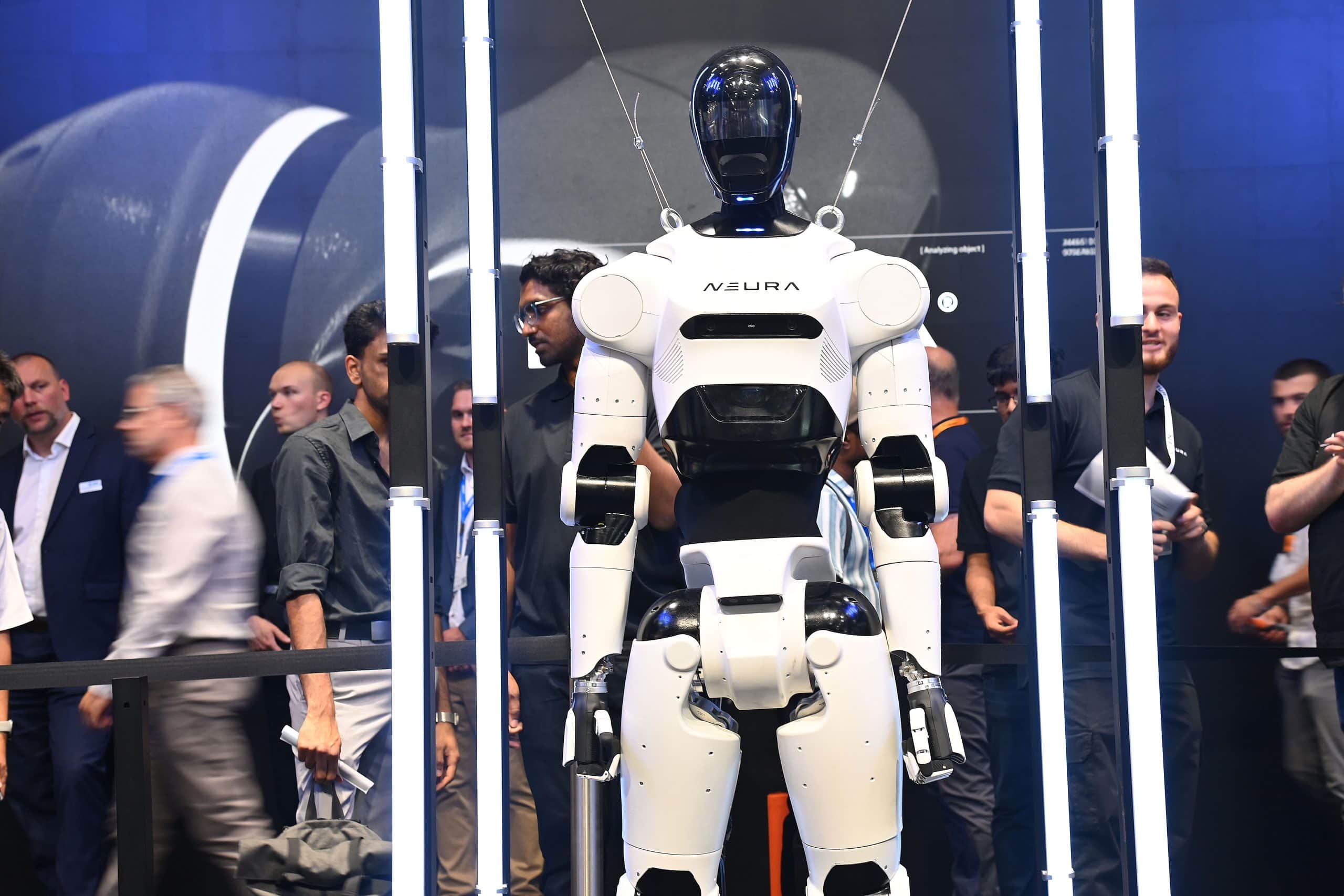 Ein humanoider Roboter mit der Bezeichnung NEURA wird auf einer Veranstaltung ausgestellt, angebunden an ein Gerät in der Nähe, umgeben von Teilnehmern und hellen vertikalen Lichtern.