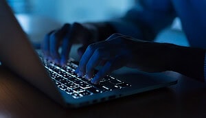 Nahaufnahme von Händen, die in einer schwach beleuchteten Umgebung auf einer Laptoptastatur tippen, wobei die Tasten durch das Leuchten des Bildschirms erhellt werden - eine Atmosphäre, die oft mit der Untersuchung von Pig-Butchering-Betrug USA-Fällen in Verbindung gebracht wird.