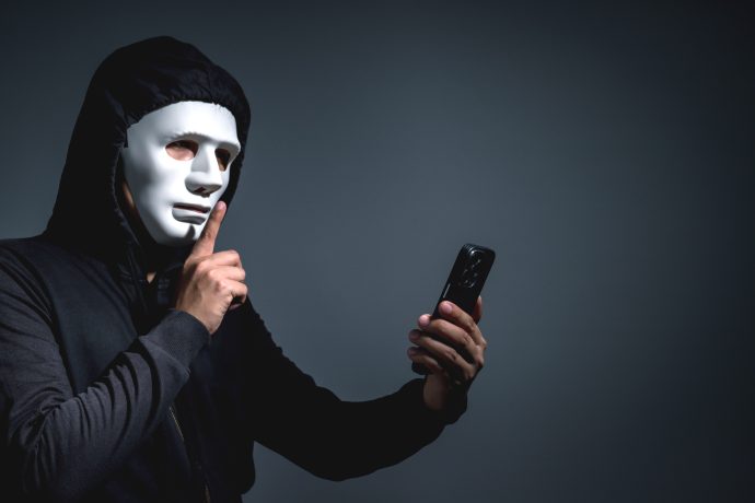 Eine Person mit schwarzem Kapuzenpulli und weißer Maske hält ein Smartphone in der einen Hand und macht mit der anderen eine "Shush"-Geste, eine subtile Anspielung auf den Fokus von ZCash auf Privatsphäre.