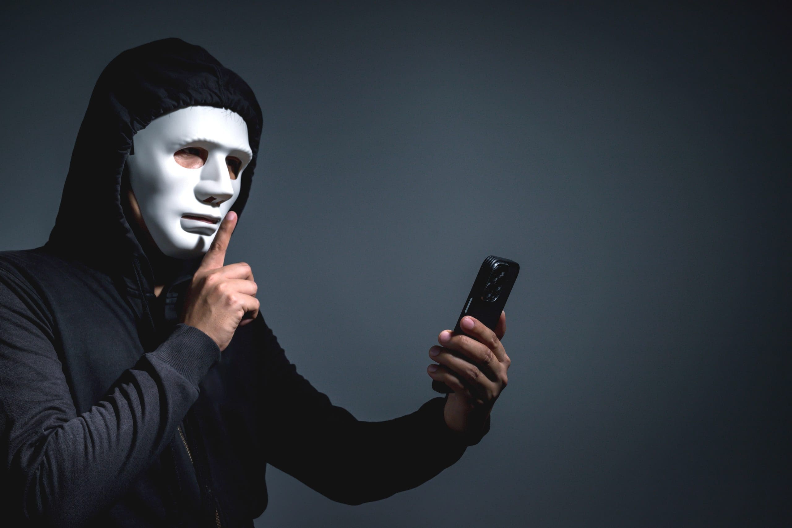 Eine Person mit schwarzem Kapuzenpulli und weißer Maske hält ein Smartphone in der einen Hand und macht mit der anderen eine "Shush"-Geste, eine subtile Anspielung auf den Fokus von ZCash auf Privatsphäre.