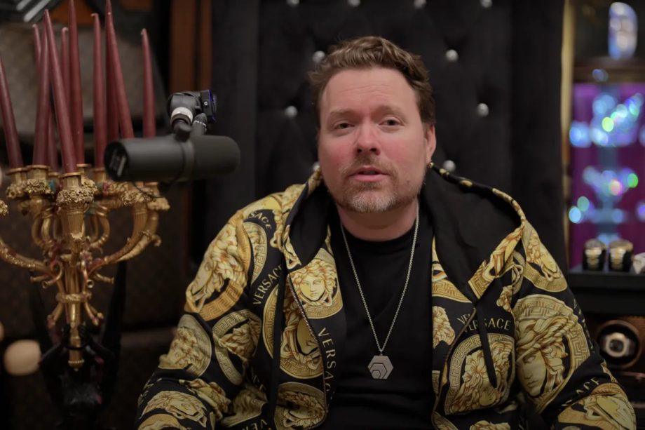 Ein Mann in einer goldenen Versace-Jacke sitzt vor einem Mikrofon, im Hintergrund stehen Kerzen und eine Vitrine, die der Szene eine rätselhafte Hex-Atmosphäre verleihen.