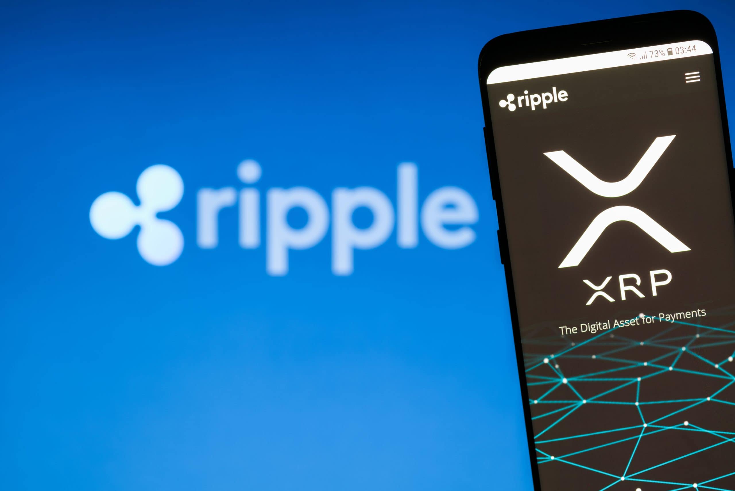 Ein Smartphone zeigt die Website und das Logo von Ripple (XRP) an. In der Ferne ist ein verschwommenes Ripple-Logo auf blauem Hintergrund zu sehen.