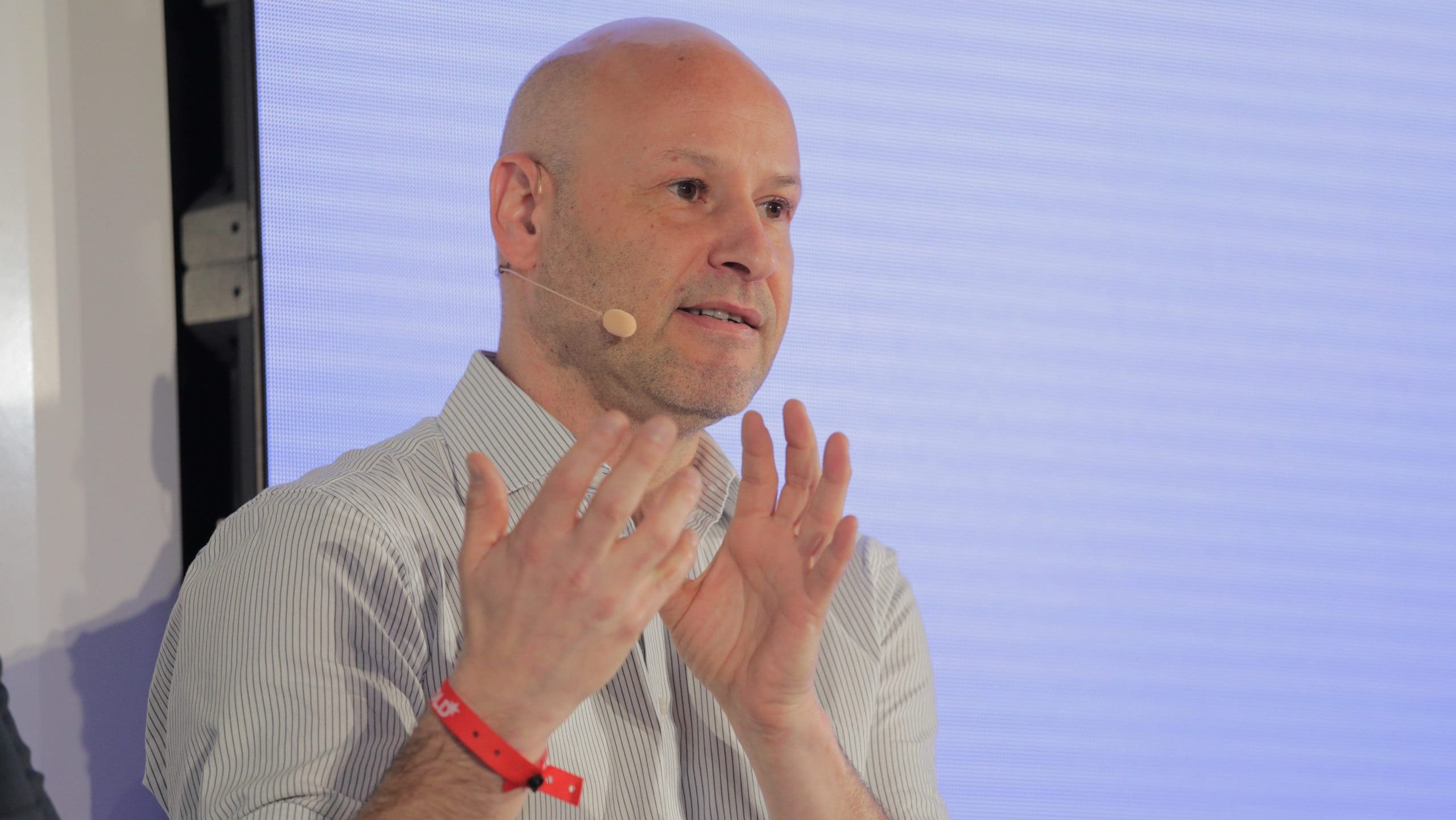 Ein glatzköpfiger Mann mit gestreiftem Hemd und Headset-Mikrofon gestikuliert mit den Händen, während er vor einem hellen Hintergrund spricht und eine ansprechende Sharplink-Präsentation hält.