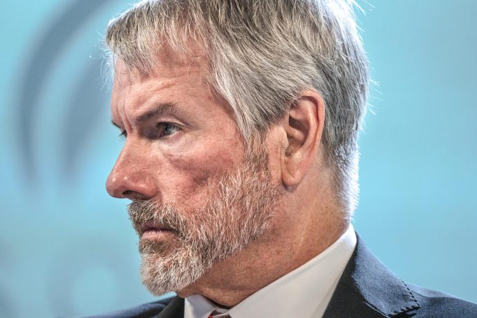 Nahaufnahme eines Mannes mittleren Alters mit grauen Haaren und Bart, der Anzug und Krawatte trägt und vor einem unscharfen Hintergrund nach links blickt und über die Auswirkungen von Bitcoin nachdenkt.