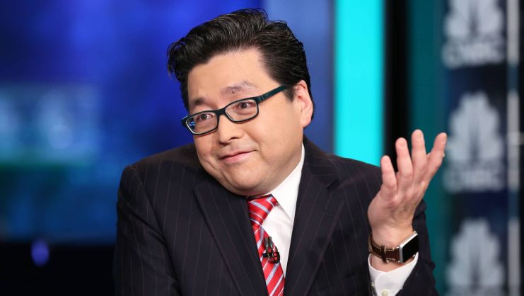 Tom Lee im Nadelstreifenanzug und mit rot gestreifter Krawatte zuckt mit einer erhobenen Hand mit den Schultern, während im Hintergrund verschwommene Bildschirme zu sehen sind, die möglicherweise Ethereum verfolgen.