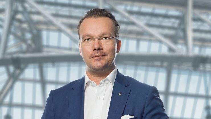 venionaire-startet-onchain-index Ein Mann in blauem Anzug und weißem Hemd steht in einem Innenraum vor großen Glasfenstern und Metallträgern.