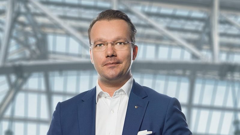 Ein Mann in blauem Anzug und weißem Hemd steht in einem Innenraum vor großen Glasfenstern und Metallträgern.