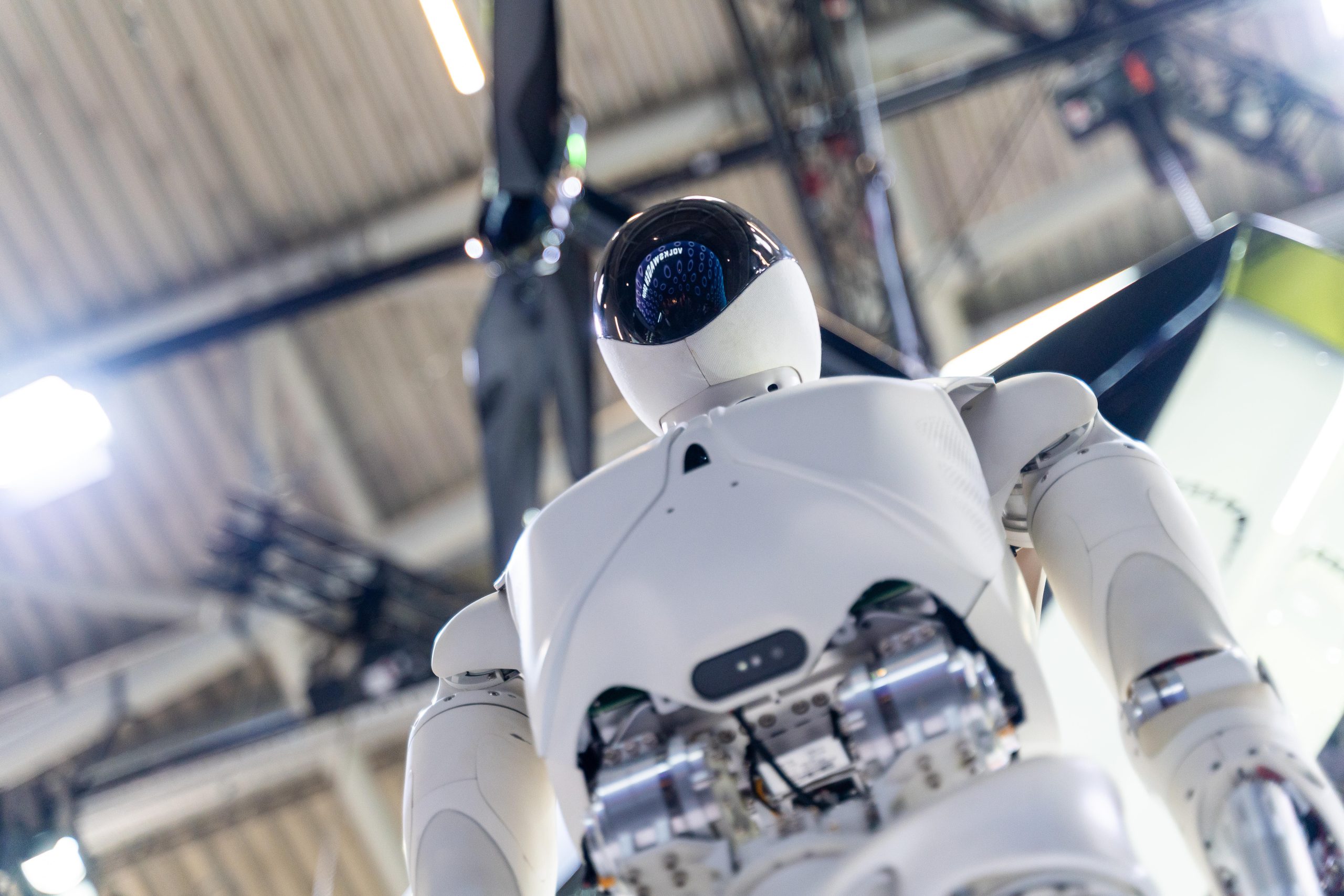 Ein humanoider Roboter in einem weißen Gehäuse mit dem Namen Krypto wird in einem Innenraum ausgestellt und zeigt seine mechanischen Gelenke und Komponenten vor einem verschwommenen industriellen Hintergrund.