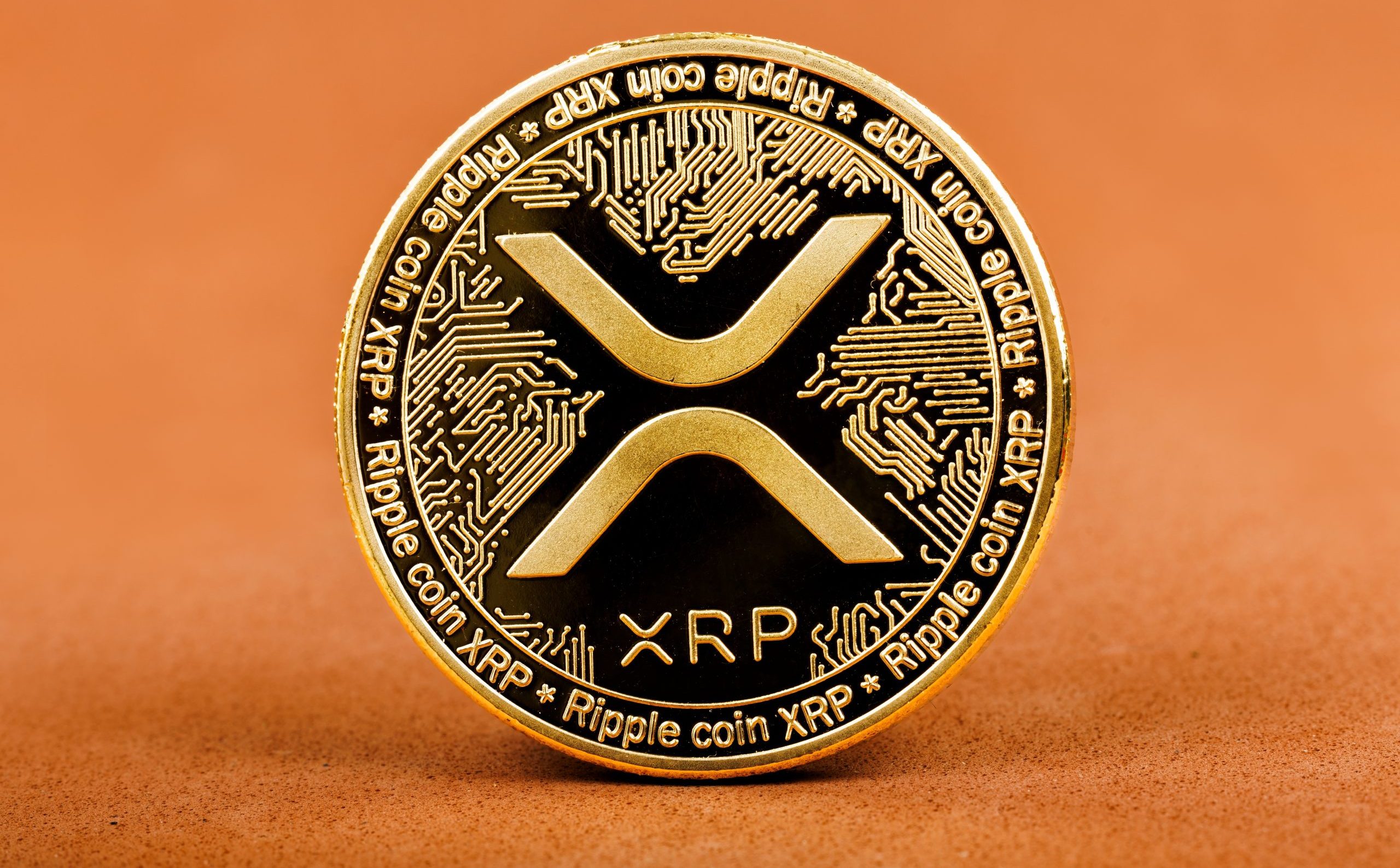 Eine goldene XRP-Münze steht aufrecht vor einem orangefarbenen Hintergrund, auf dem das Logo und kreisförmige Muster zu sehen sind.
